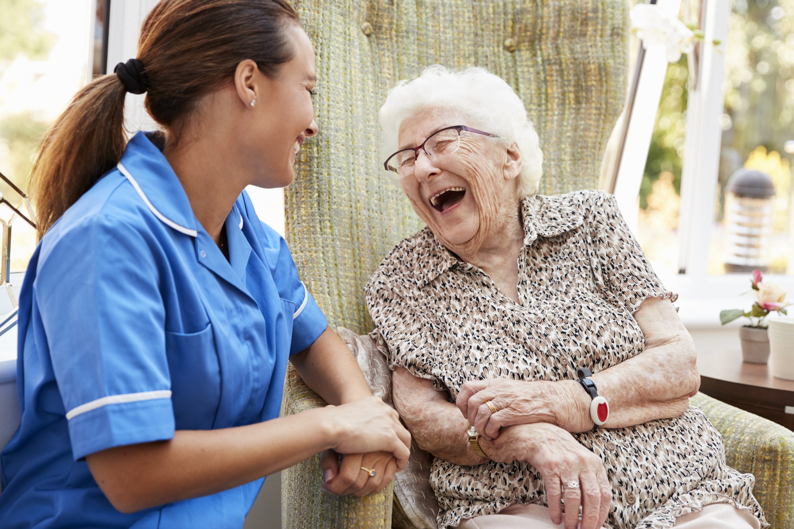 Care worker in blue uniform holding hands with elderly woman with white hair and glasses, both laughing together on floral armchair