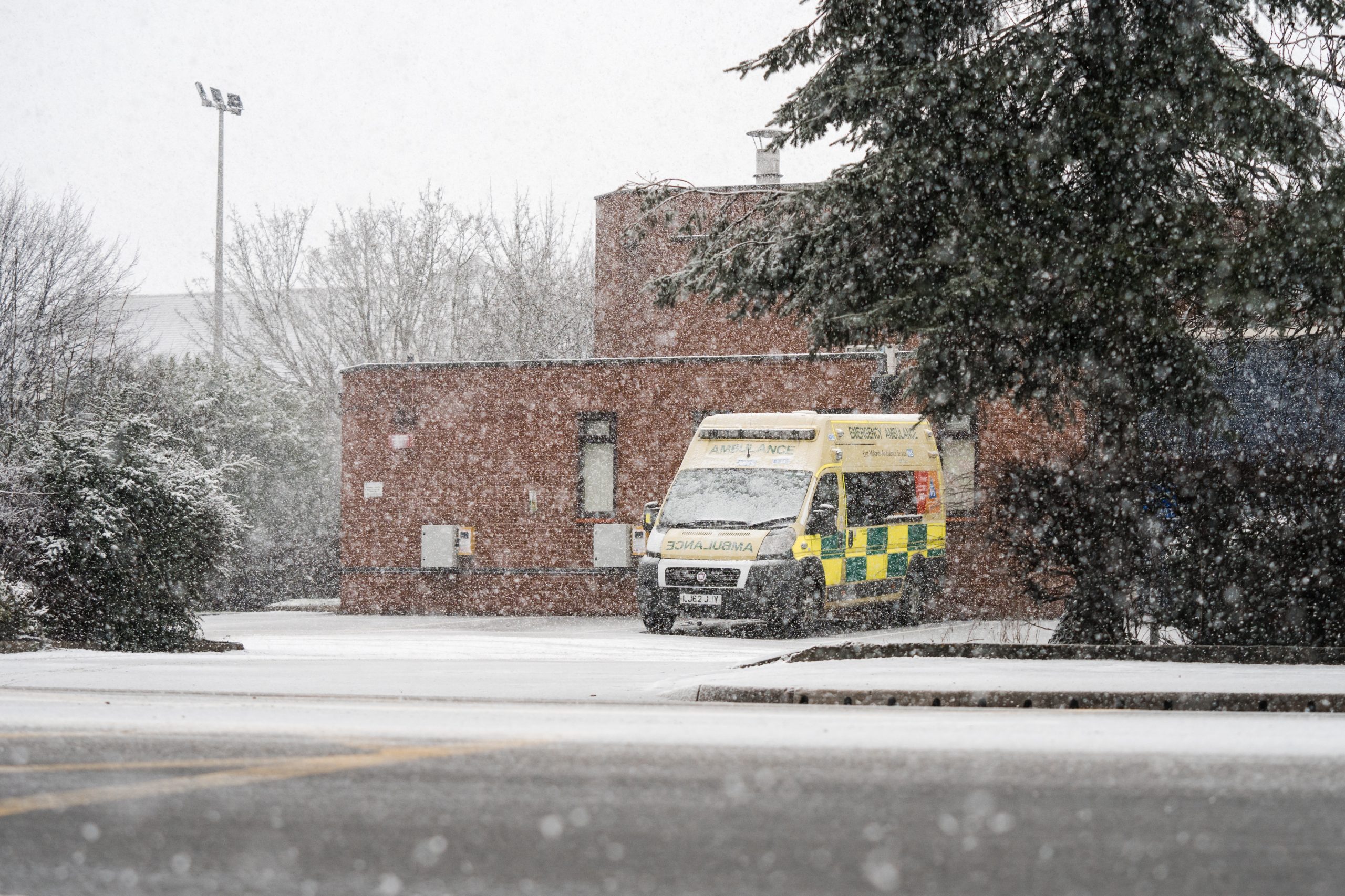 A winter scene showing an ambulance parked in a snowy hospital car park with a tree in the foreground