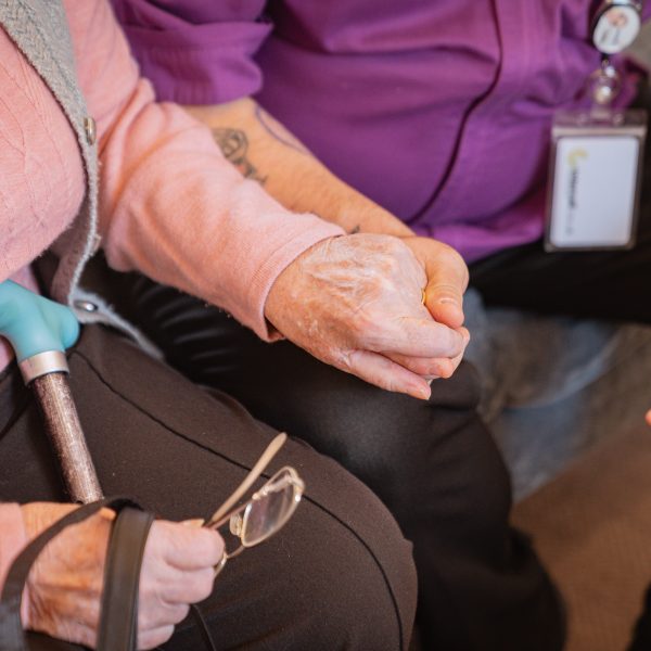 Healthcare worker in purple scrubs holding hands with elderly wheelchair user showing compassionate care by Blue Lozenge