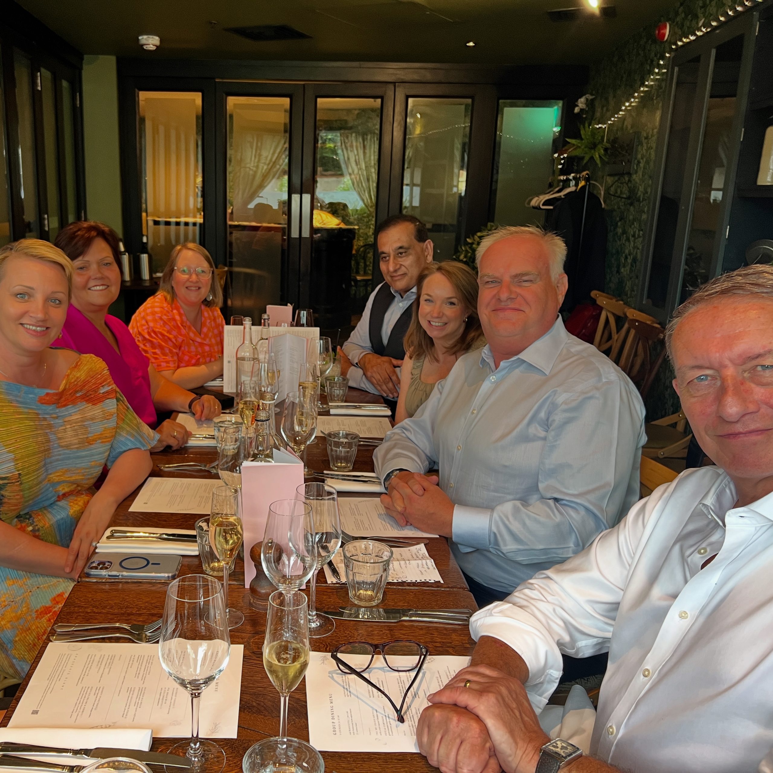 Seven people seated around restaurant table with menus, wine glasses and place settings in modern dining space