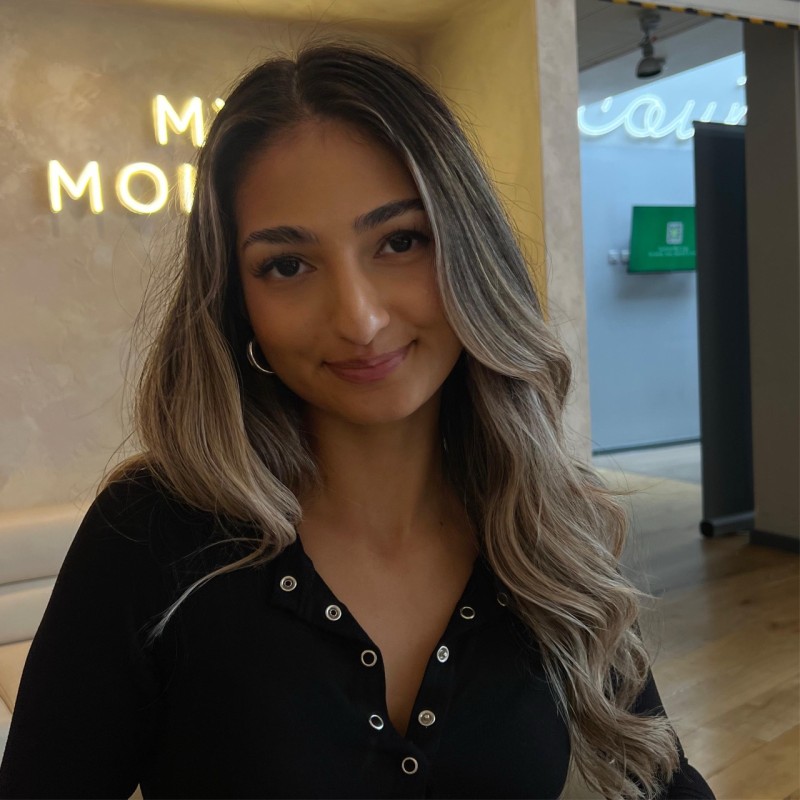 Woman smiling at the camera, with long wavy highlighted hair, wearing a black top. She is indoors with a softly lit background.